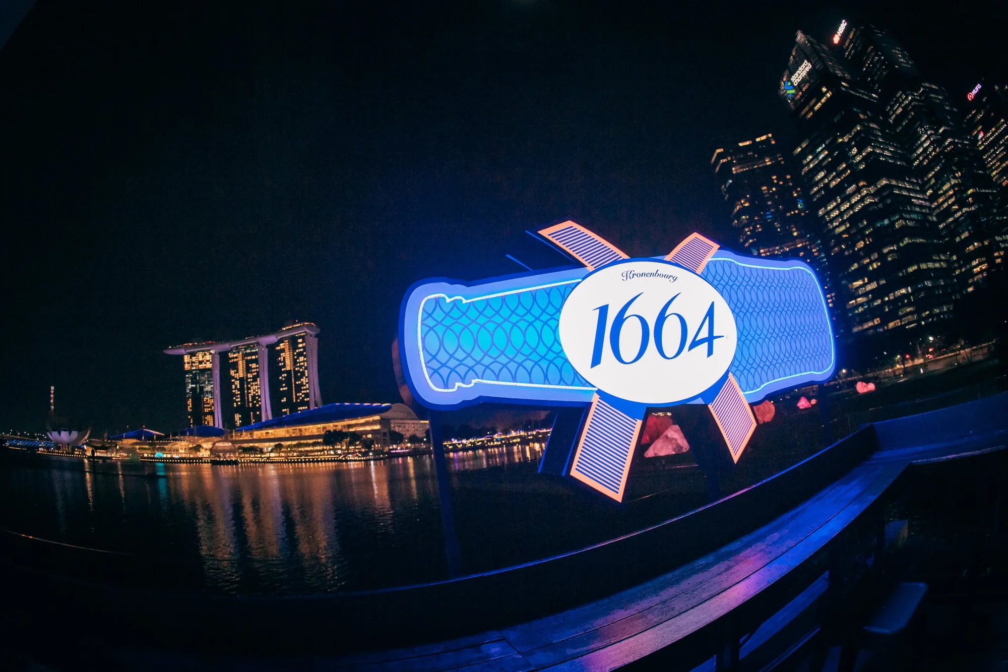 A lit 1664 sign shaped like the bottle label, with Marina Bay Sands across the water